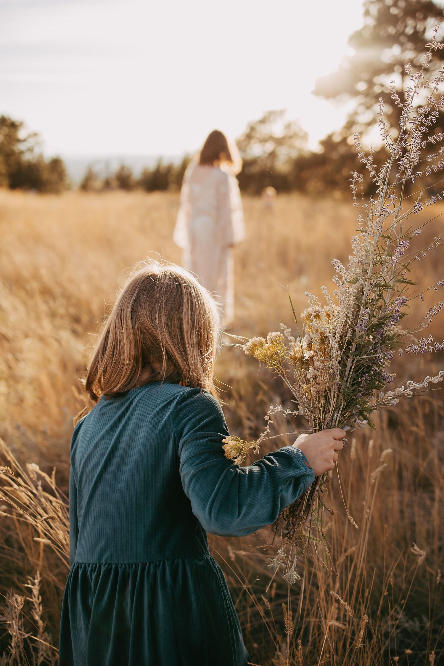 Young girl in teal dress picking wildflowers in golden field at sunset with woman in white dress in background.