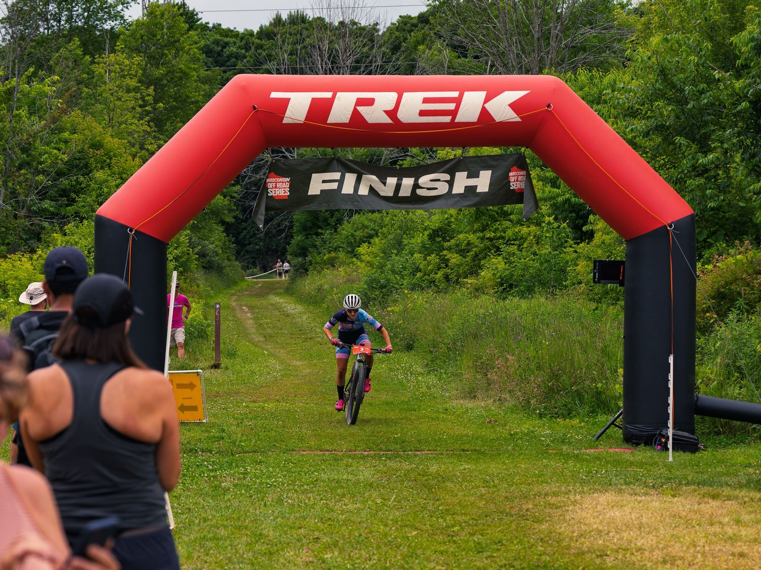 A mountain bike racer approaches the finish arch at a Wisconsin Off Road Series race, riding a grassy trail toward the red TREK banner.