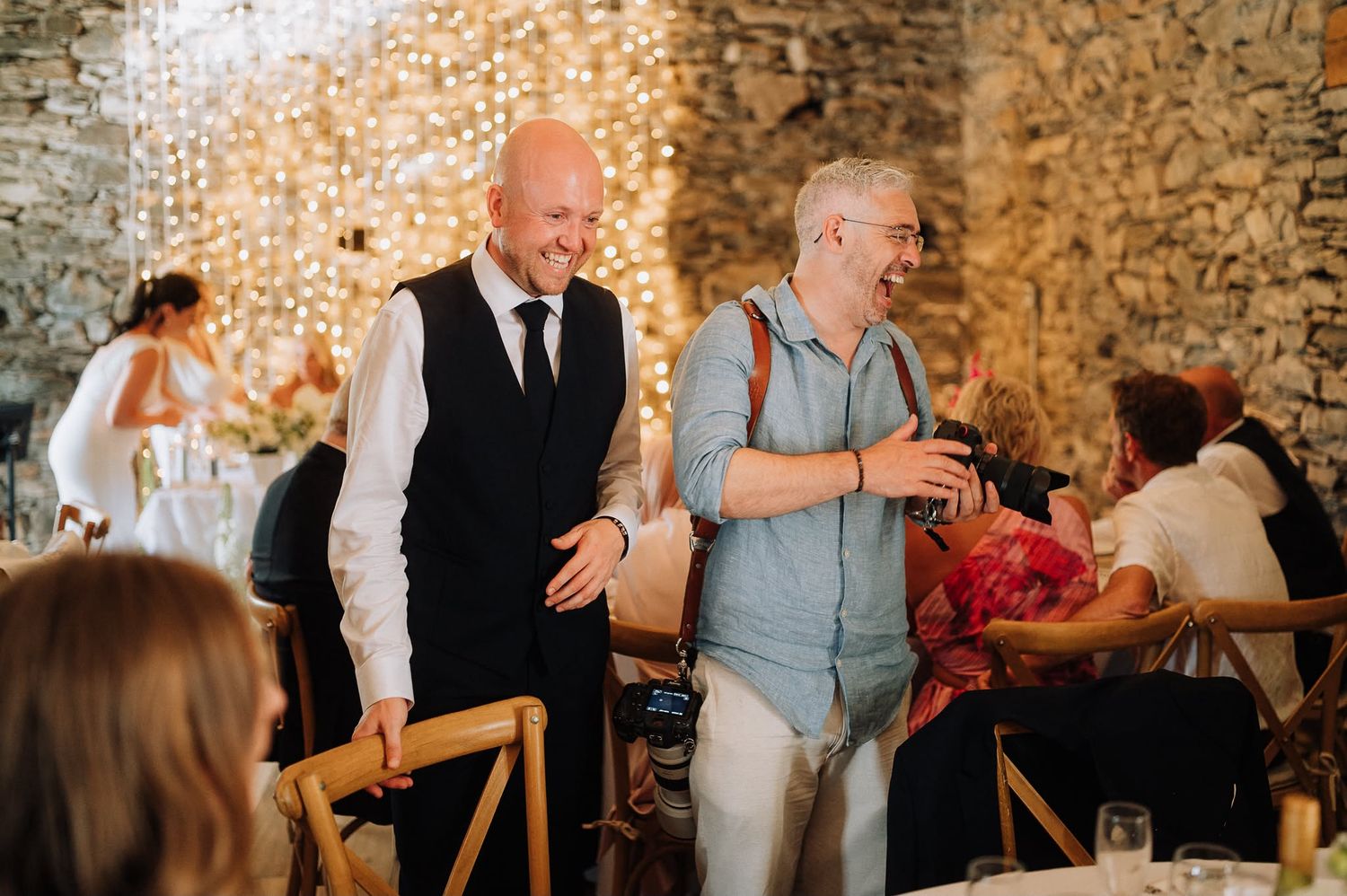 Two men laughing and shaking hands at a wedding reception in a rustic stone venue with fairy lights.