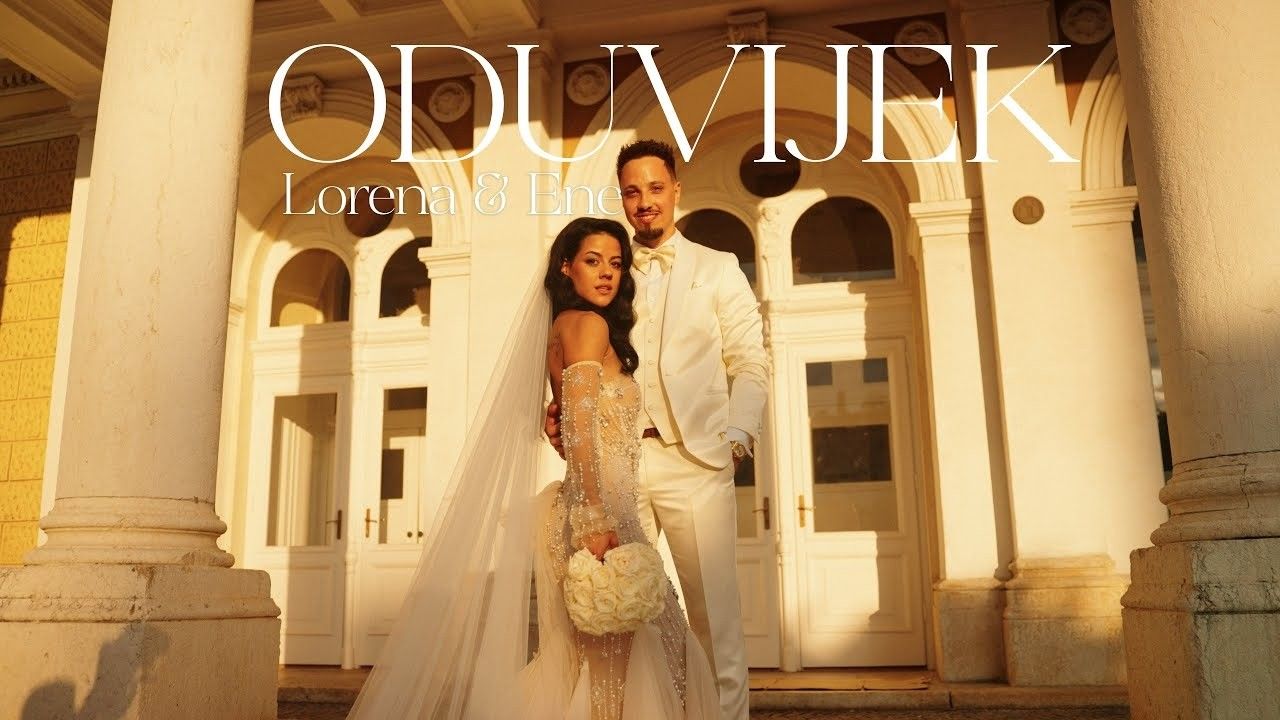 Wedding couple poses before grand columns, bride in lace gown with veil, groom in white suit, text reads ODUVIJEK.