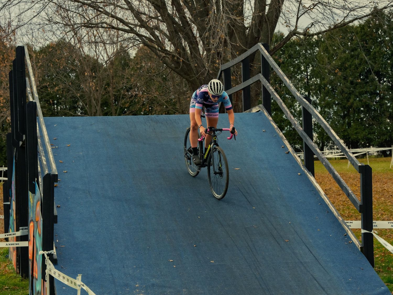 A cyclocross racer rides down a large blue wooden ramp on a TREK-taped course, coasting toward the camera under bare autumn trees.