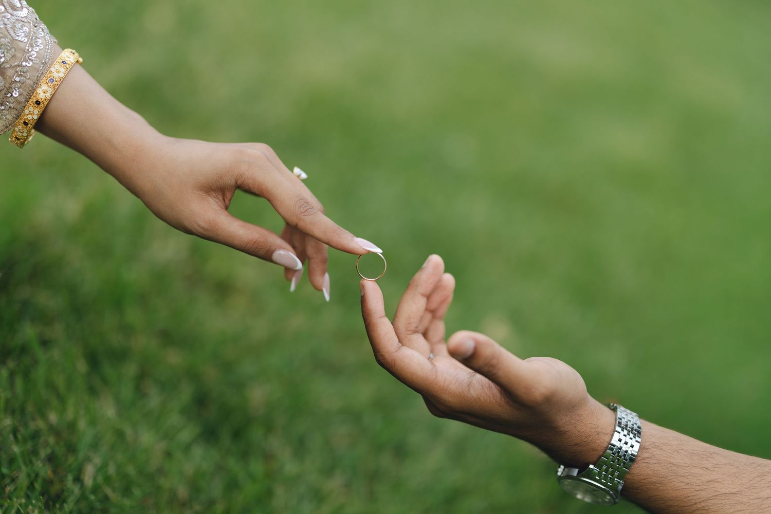 Two hands reaching toward each other over green grass, one wearing a gold bracelet with manicured nails, the other a silver watch.