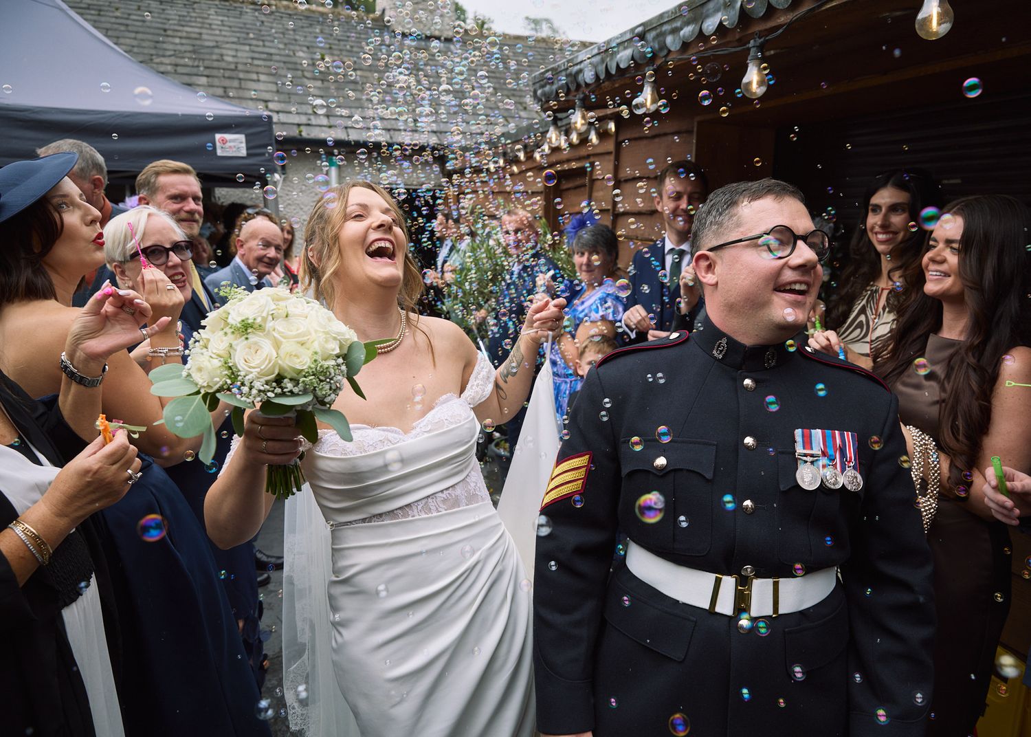 Bride in white gown and groom in military uniform celebrate with confetti and bubbles after wedding ceremony.