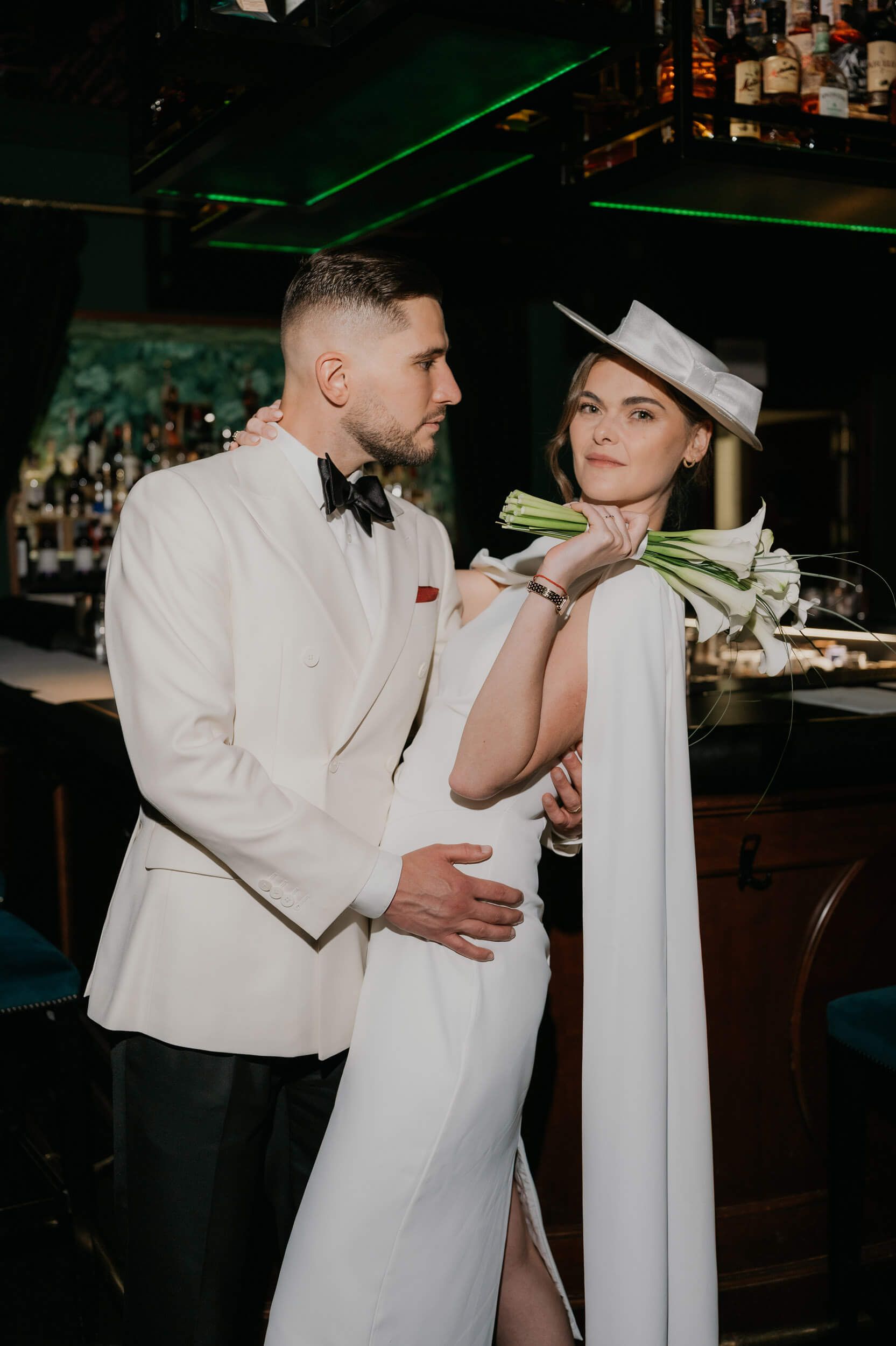 Elegant couple in white wedding attire pose at a bar, groom in tuxedo embracing bride in cape dress and hat.
