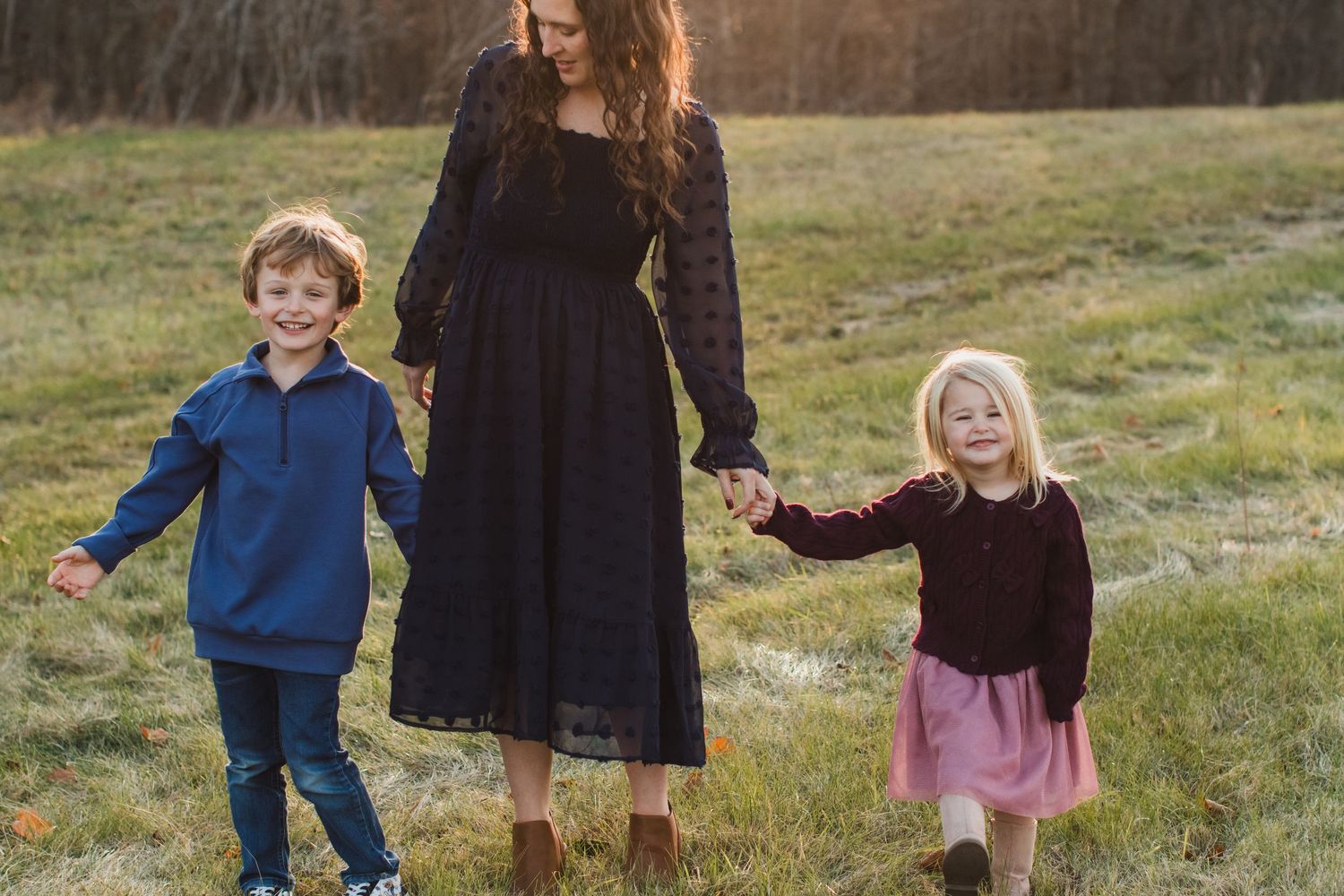 A mother in a black lace dress walks hand-in-hand with her young son and daughter in a field.