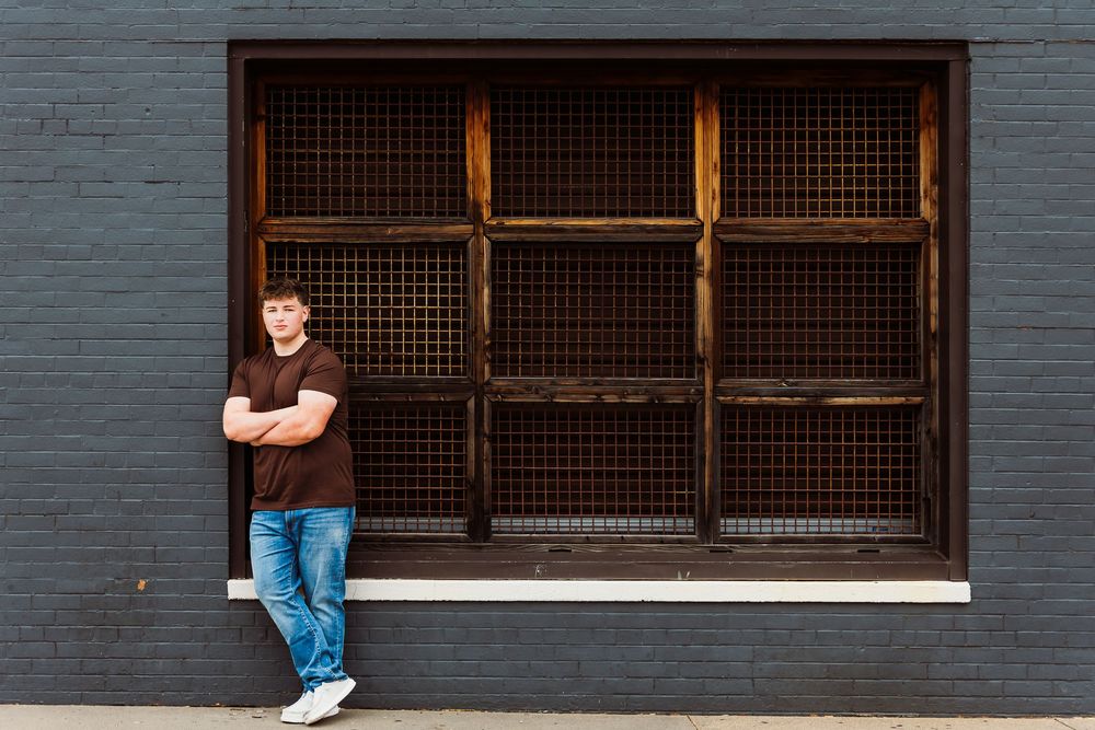 Young man in brown shirt and jeans leaning against a dark brick wall with a large industrial grid window.