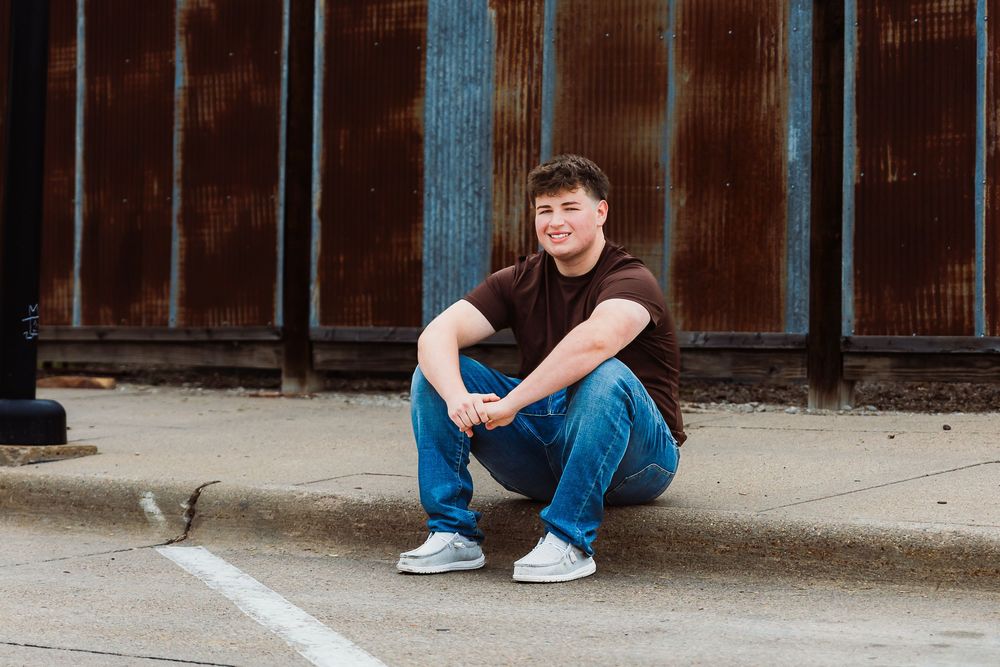 Young man in jeans and maroon shirt sitting on curb smiling, posed for senior portrait outdoors.