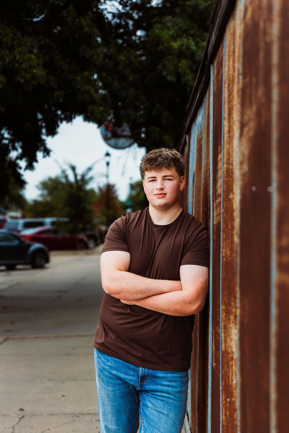 Young man in brown t-shirt and jeans leaning against rustic metal wall with arms crossed outdoors.