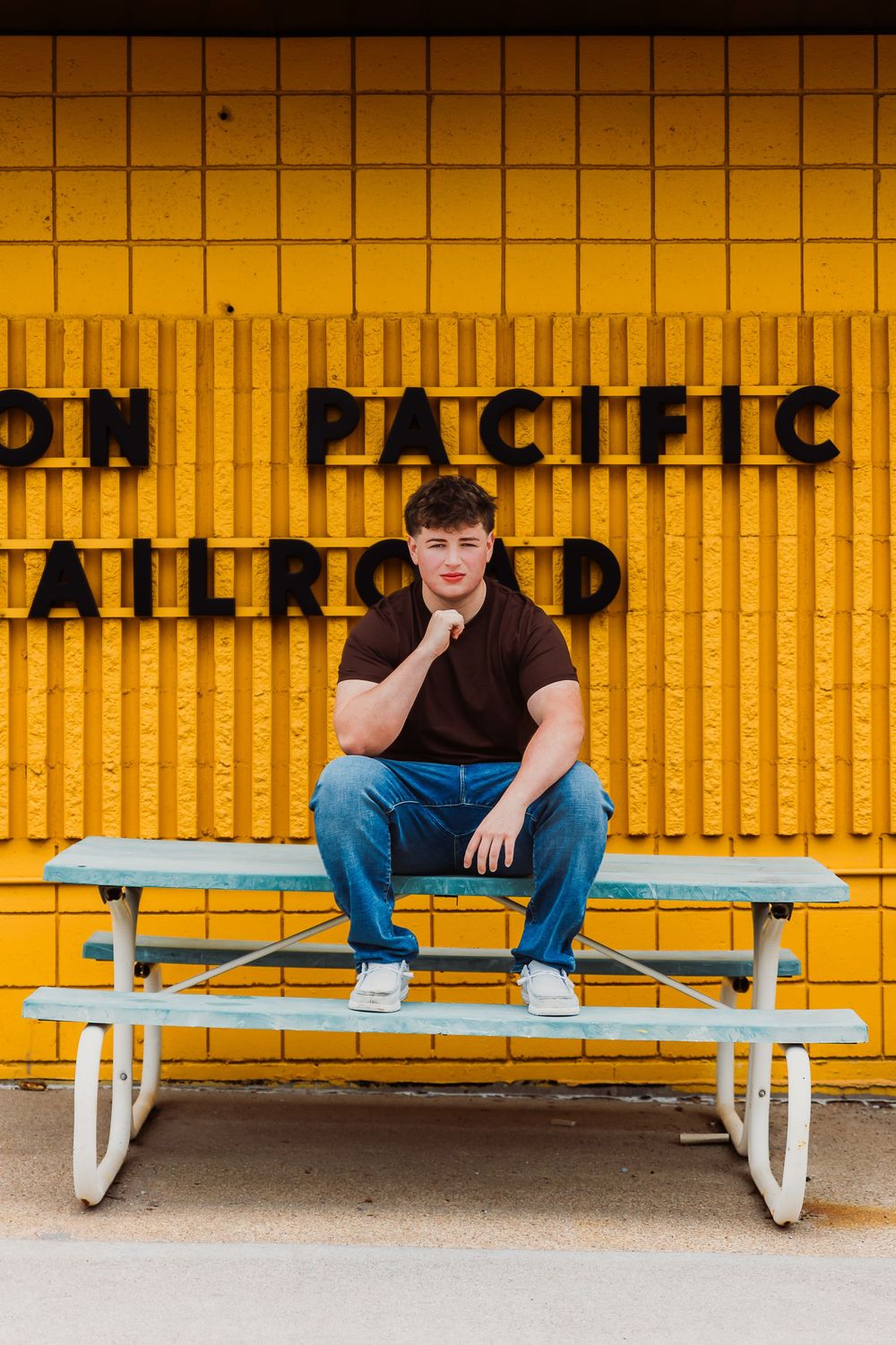 Young man in dark shirt and jeans sits on a picnic table in front of a yellow Union Pacific Railroad wall.