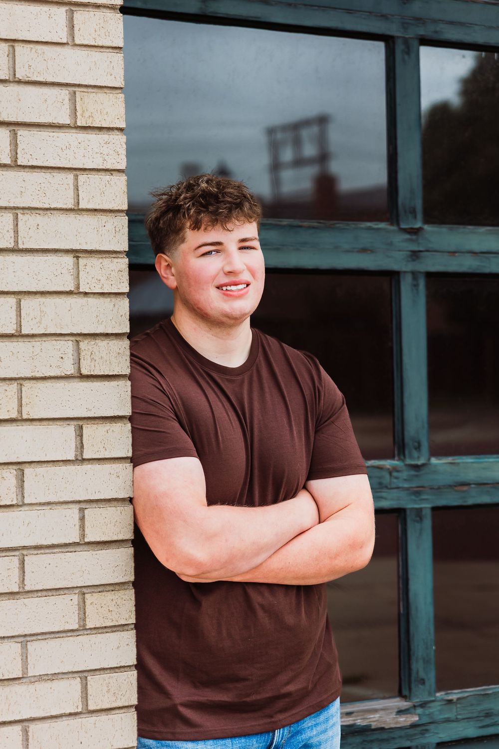 Young man in brown t-shirt leaning against brick wall with arms crossed, smiling confidently outdoors.