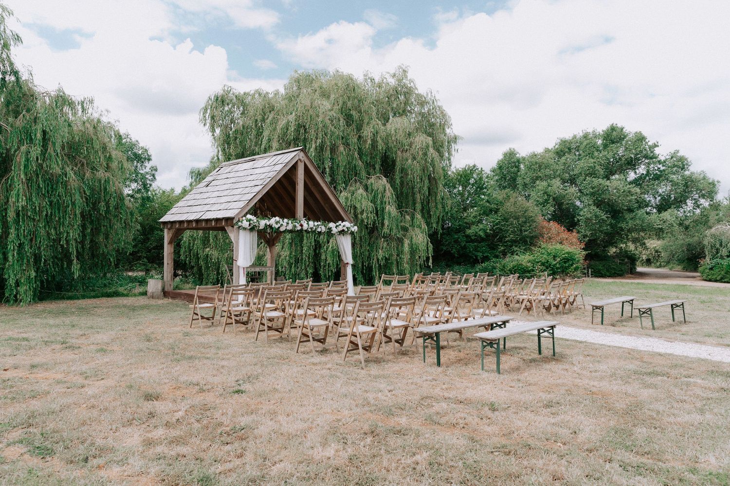 Cromwell Manor Outdoor garden Pagoda wedding ceremony setup with wooden chairs, floral-adorned pavilion, and weeping willow tree backdrop.