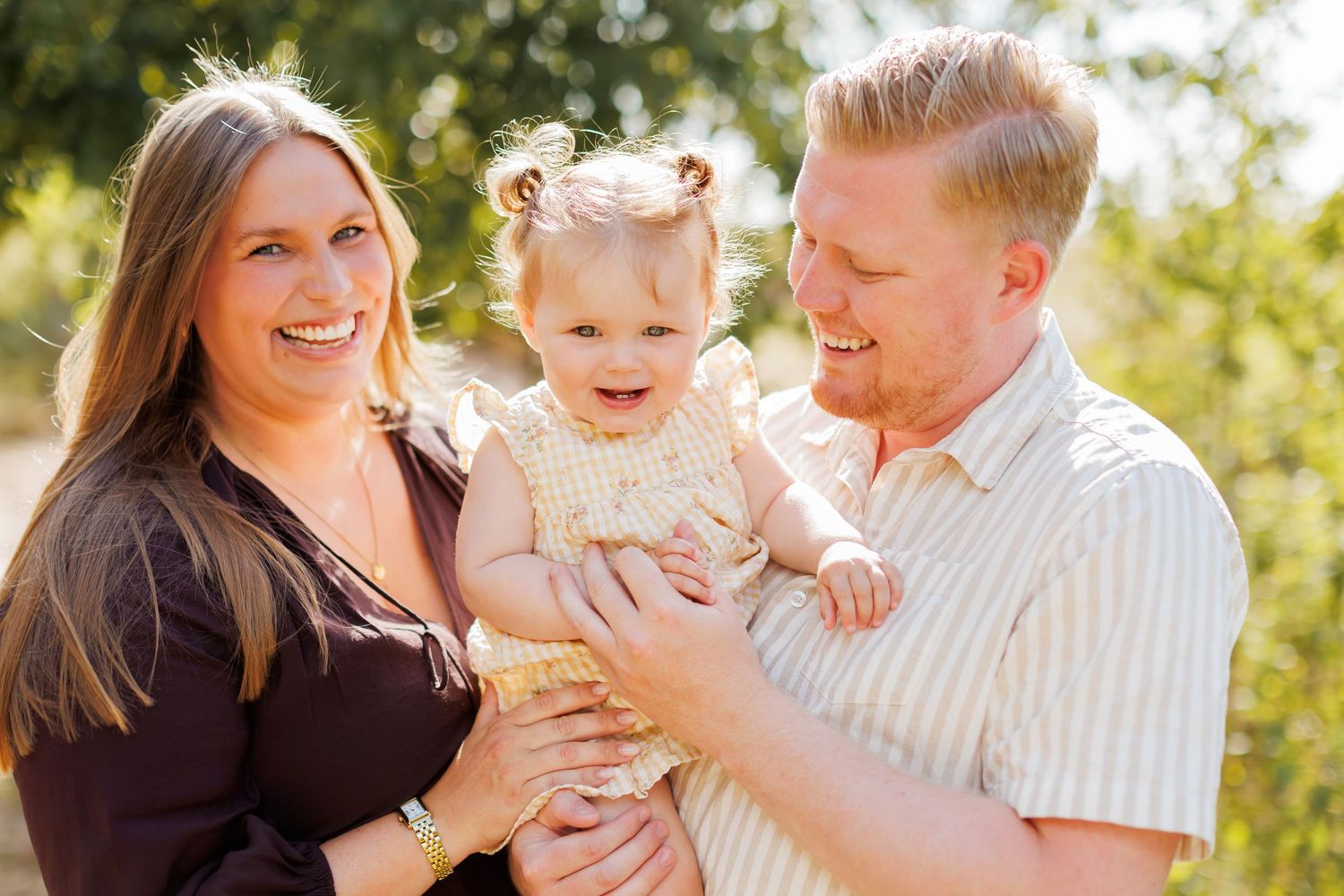 Happy family portrait with smiling parents holding toddler girl outdoors in natural sunlight.