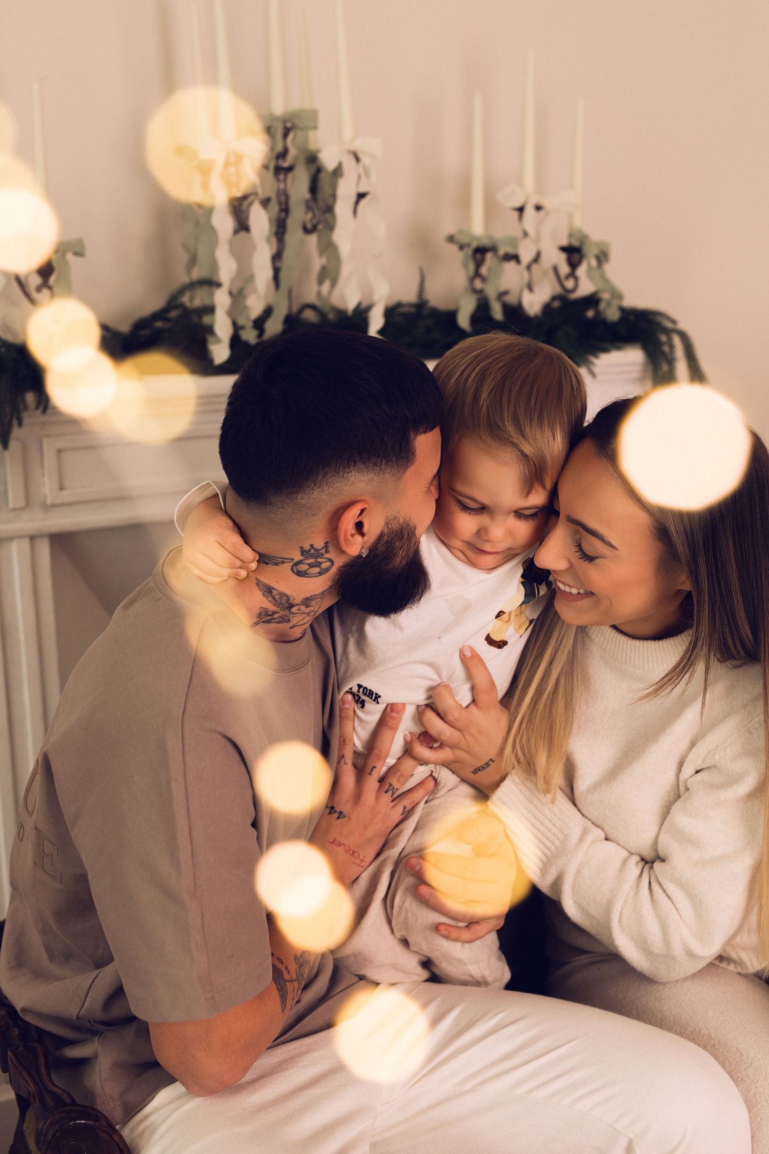 A tattooed father and smiling mother share a tender moment with their young child near a decorated fireplace.