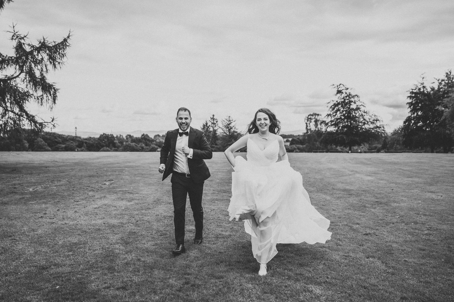 Newlyweds running happily across open field in black and white photography.