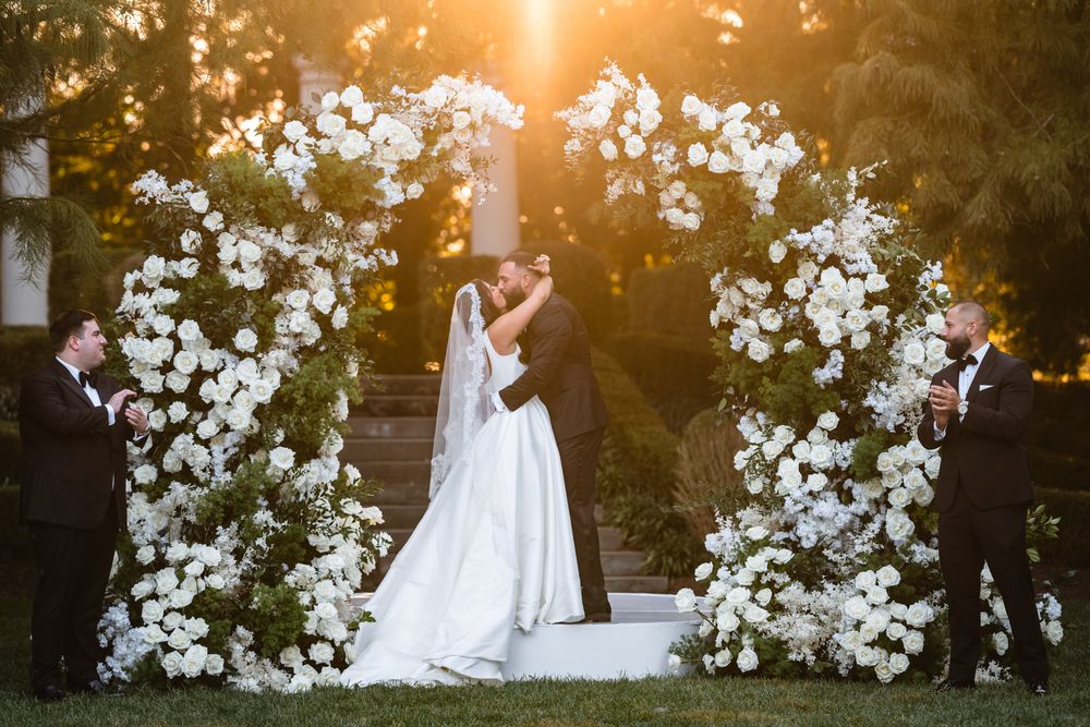 A sunset wedding ceremony under an arch adorned with white floral arrangements in a garden setting.
