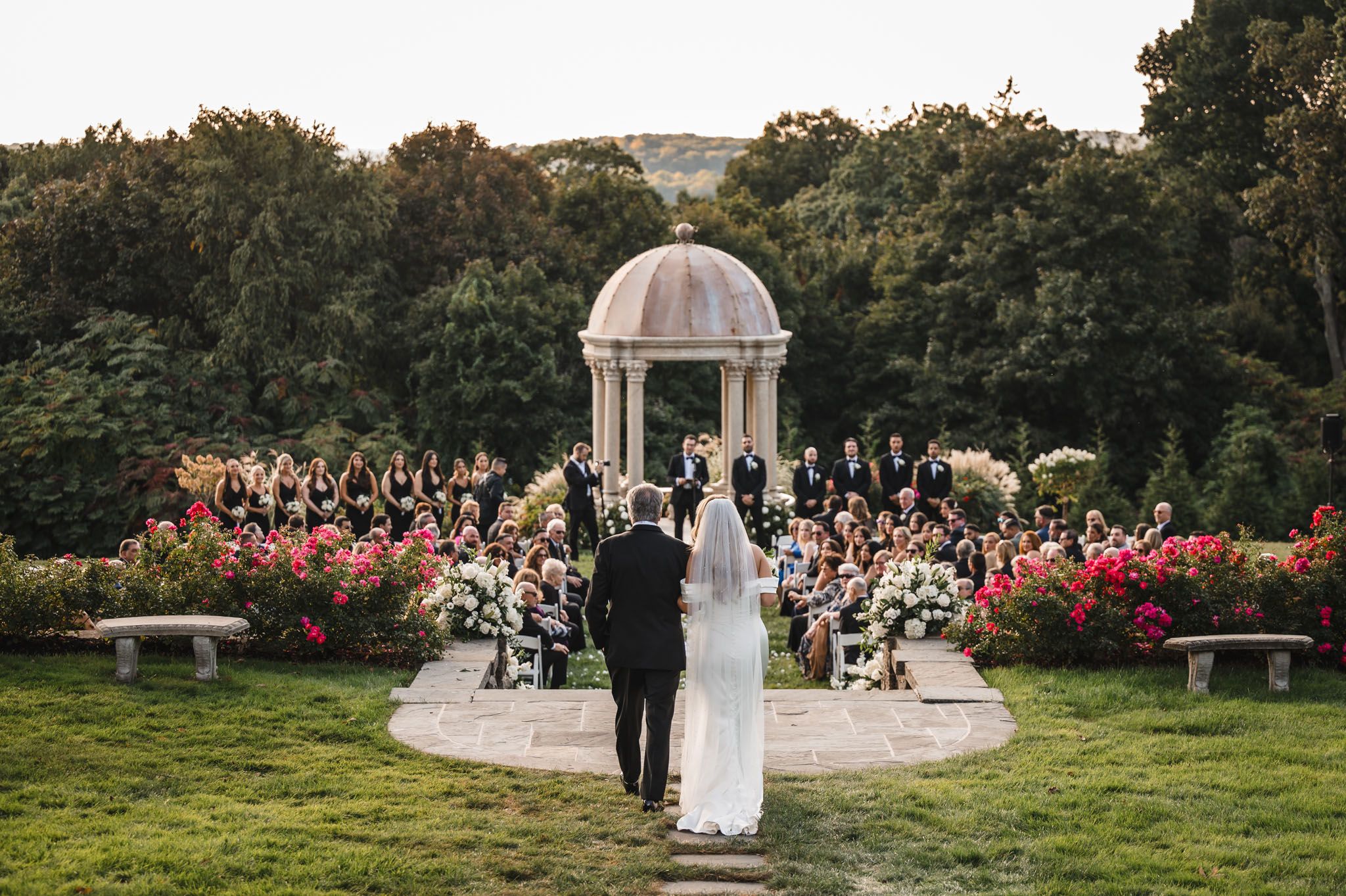 Panoramic view of an outdoor wedding ceremony setup with white chairs and gazebo surrounded by lush gardens.