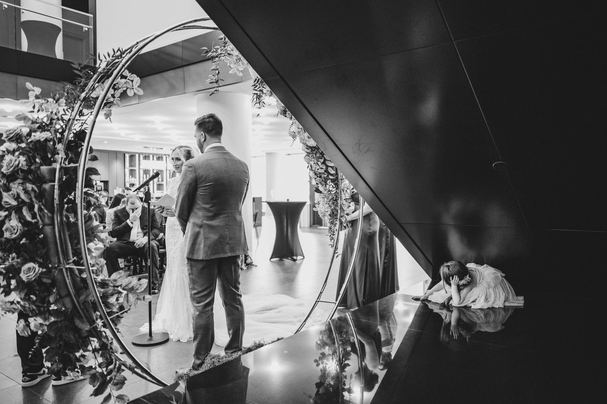 Black and white photo showing a wedding ceremony setup with a circular arch and floral decorations indoors.
