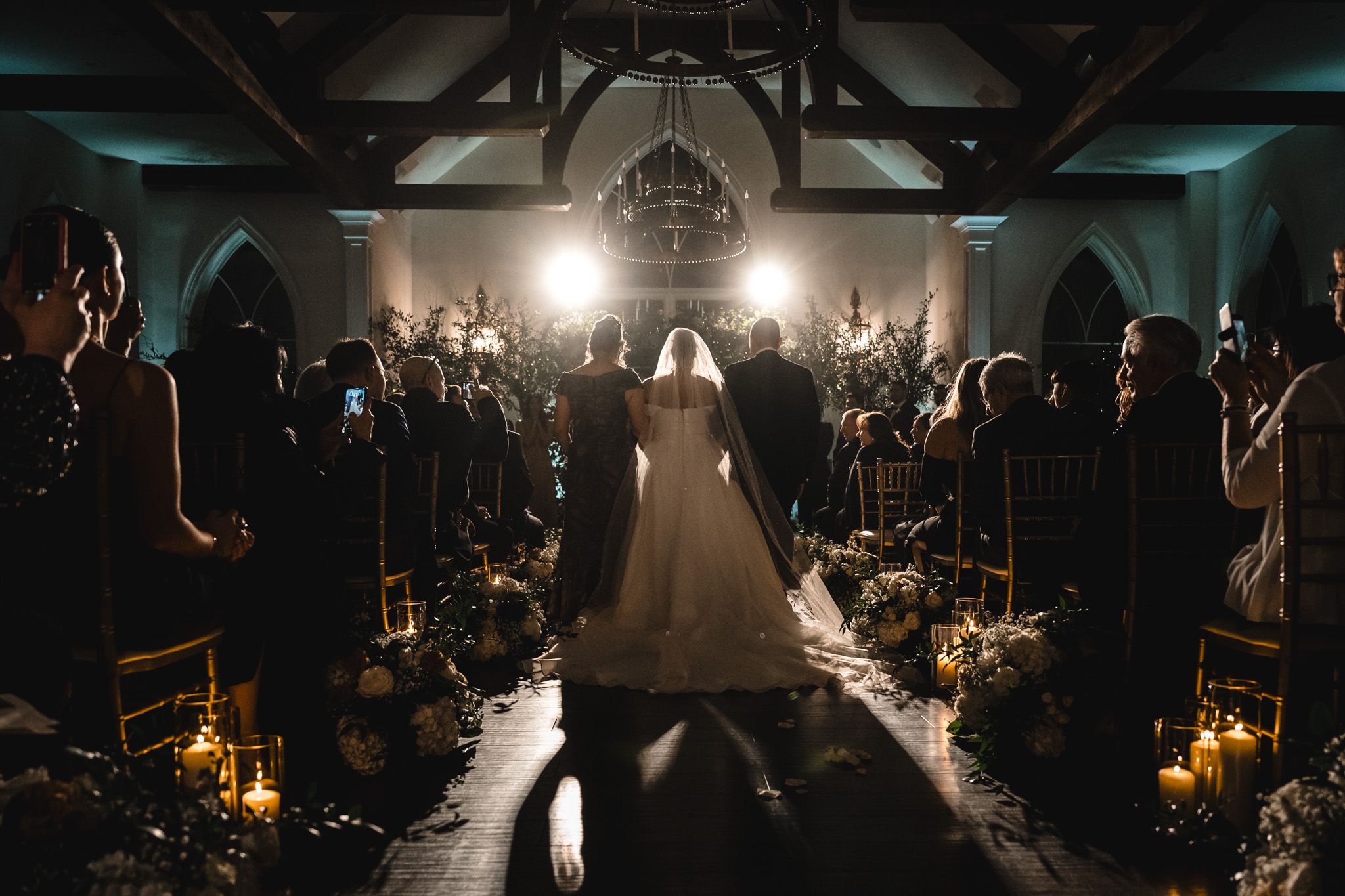 Silhouetted wedding couple walks down a dramatically lit church aisle with guests seated on both sides.