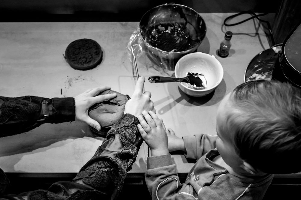 Black and white overhead shot of adults and child working together at a kitchen table with bowls and baking ingredients.