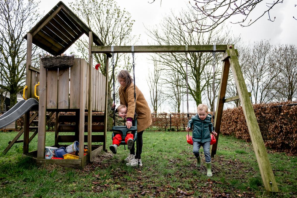 A mother pushes two young children on swings in a wooden backyard playset with slide on a cloudy day.