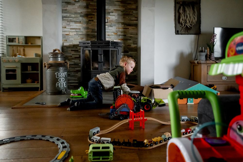 Young boy playing with toy trains and vehicles on a wooden floor in a cozy living room with a wood stove.