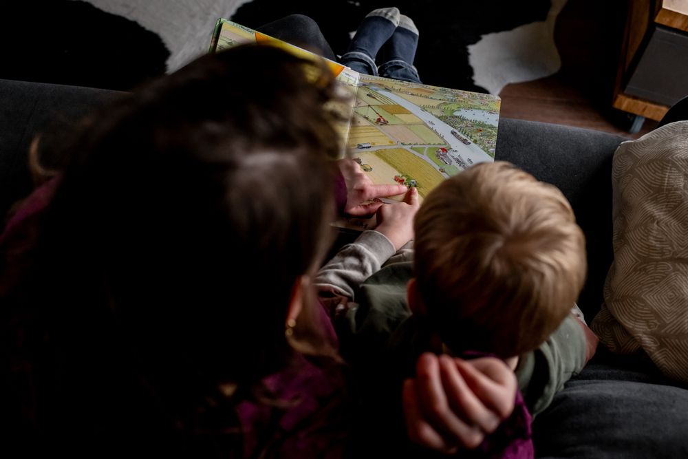 Two children cuddle together on a couch reading a colorful picture book in a cozy, dimly lit room.