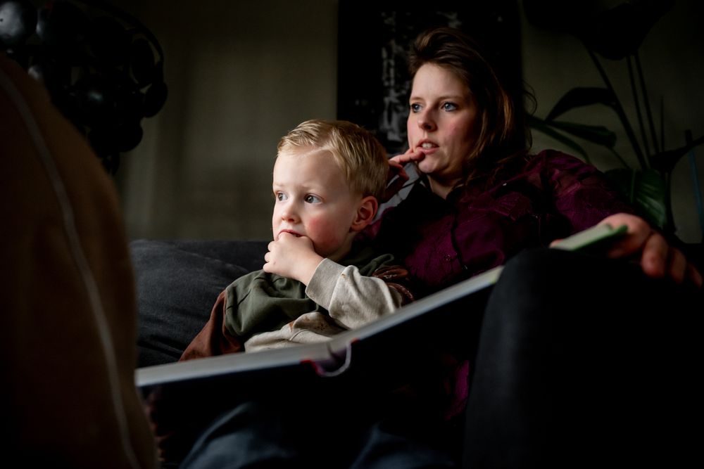 Woman and young child sitting on couch together reading a book in a dimly lit room.