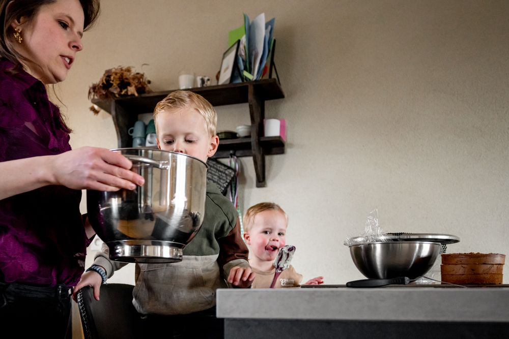 Mother baking with two young children in kitchen, toddler smiling near mixing bowls on counter.