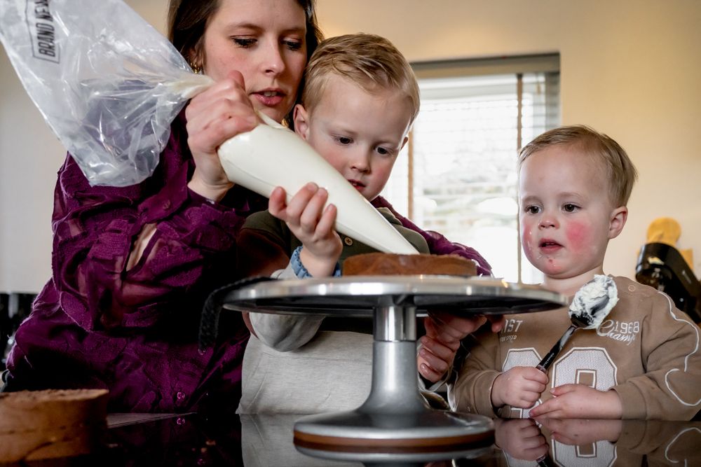 A mother and two young children decorating a cake together using a piping bag in a cozy kitchen.