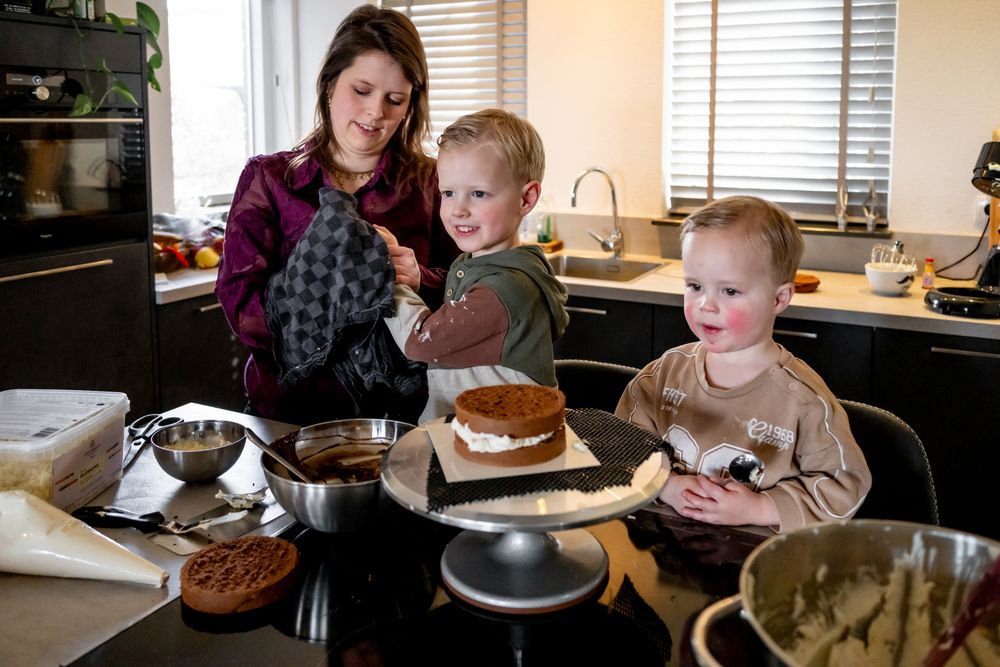 A mother and two young children baking a layered cake together in a modern kitchen with baking supplies on the counter.