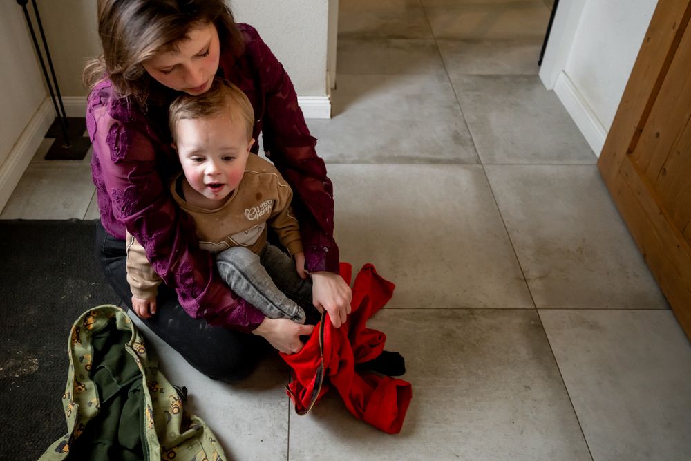 Mother sitting on tile floor helping toddler with red jacket.