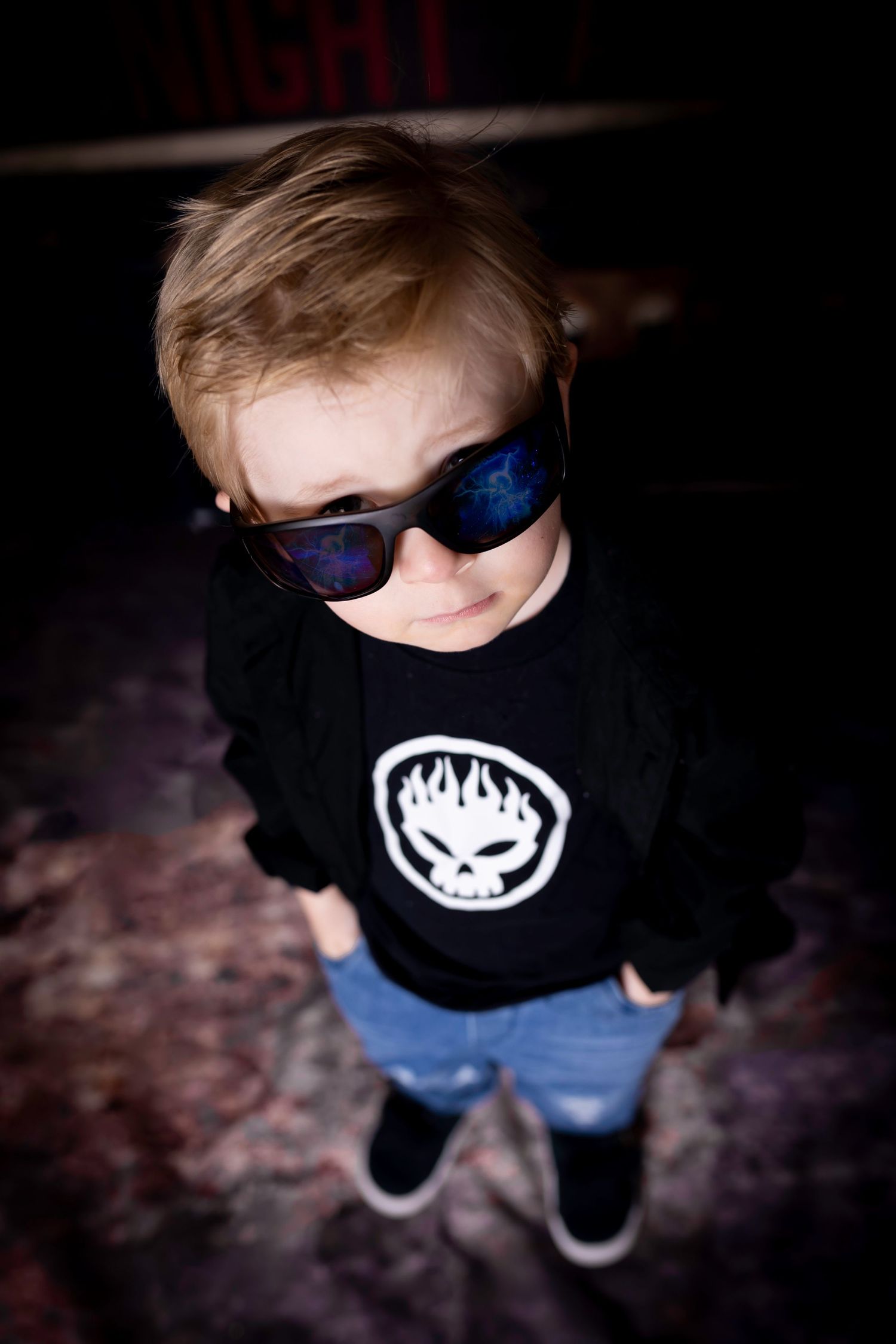 Cool kid in black t-shirt with logo and blue sunglasses standing against dark background shot from above.