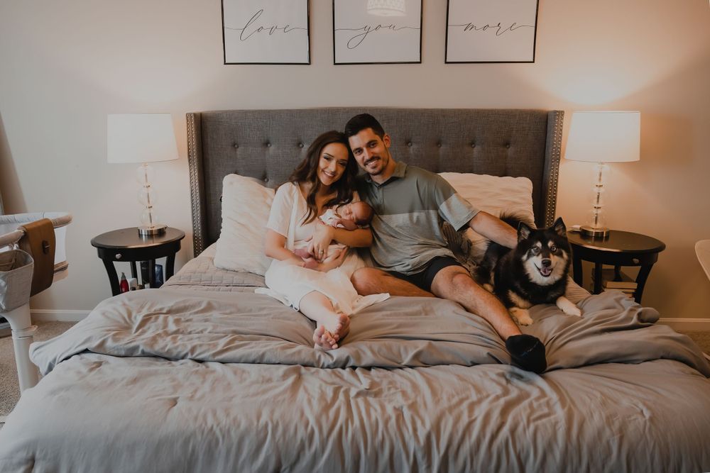 Family lounging on gray bed with newborn baby and pet dog.