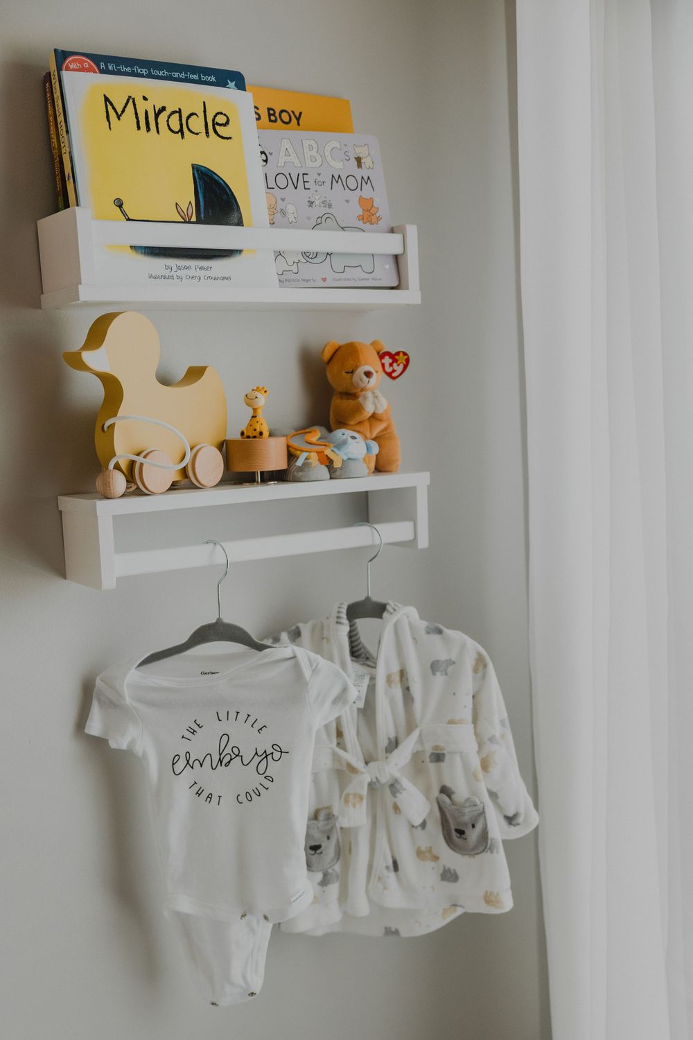 Nursery shelf displaying children's books, stuffed animals, and baby clothes hanging below.