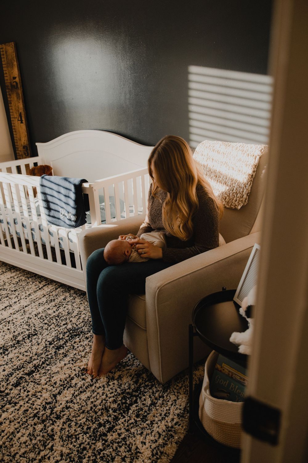 A person sits in a cozy nursery chair next to a white crib with natural light streaming through blinds.