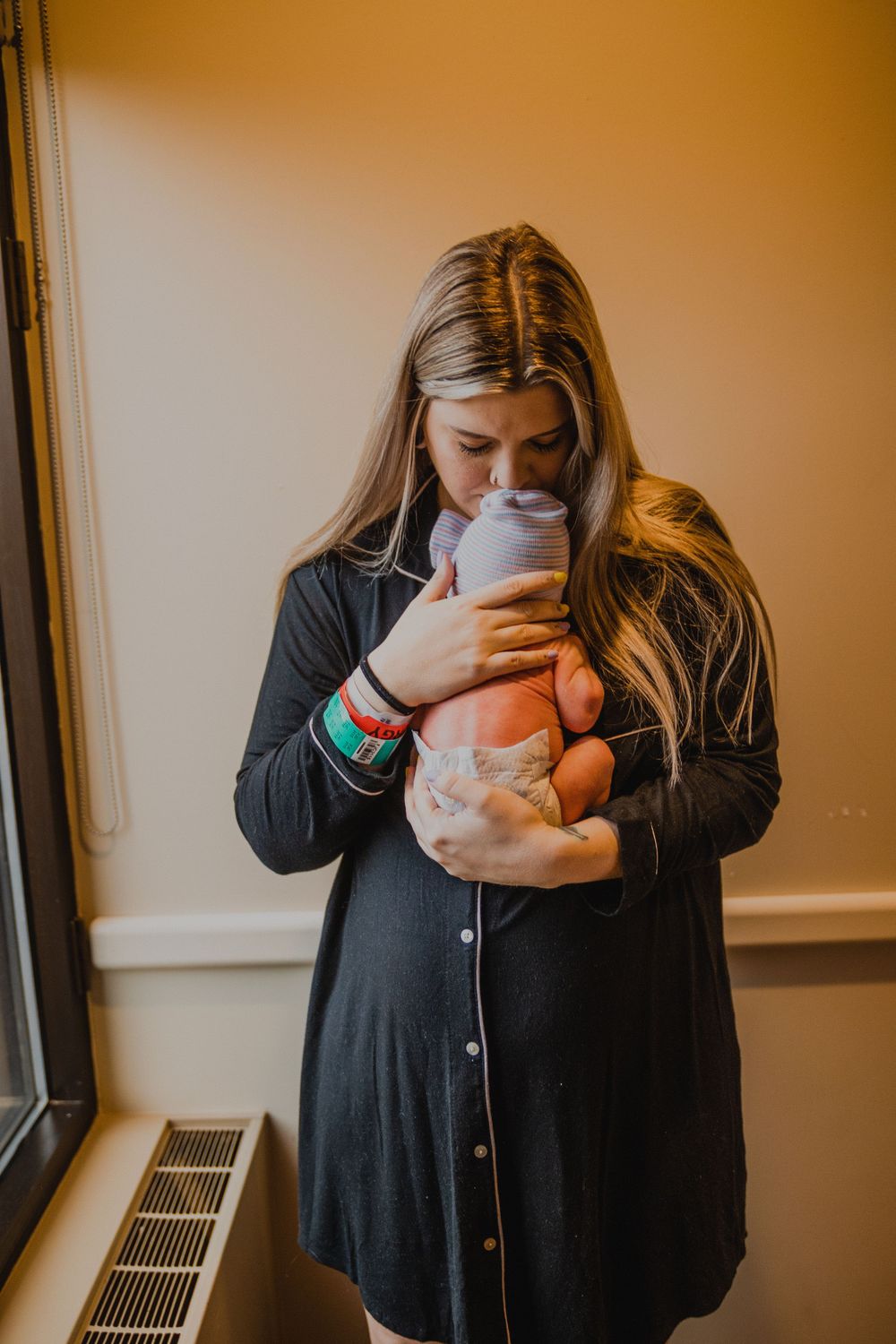 Person in a black cardigan tenderly holding their newborn baby near a window.