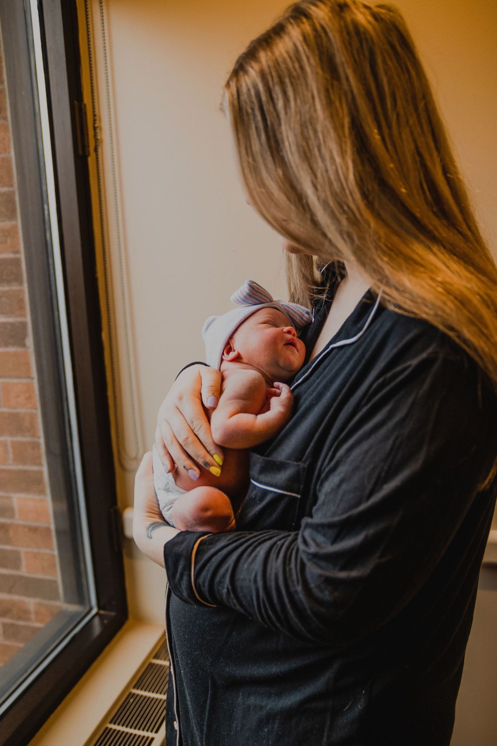 Parent in dark clothing holds sleeping newborn while standing by window.