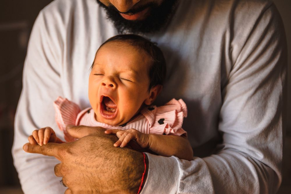 Baby yawning while being held in caregiver's arms wearing a gray sweater.