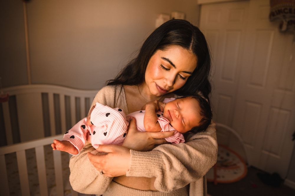 A tender moment as a new parent in a cozy beige sweater cuddles their sleeping infant next to a white crib.