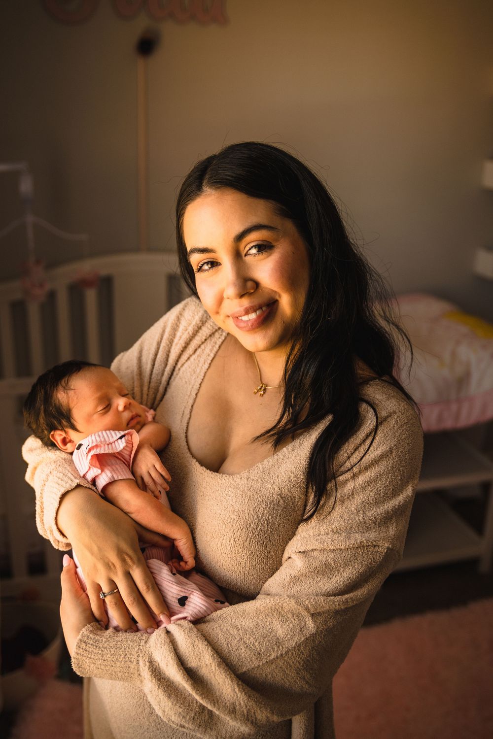 A glowing parent in tan sweater smiles while holding their newborn baby in a nursery setting.