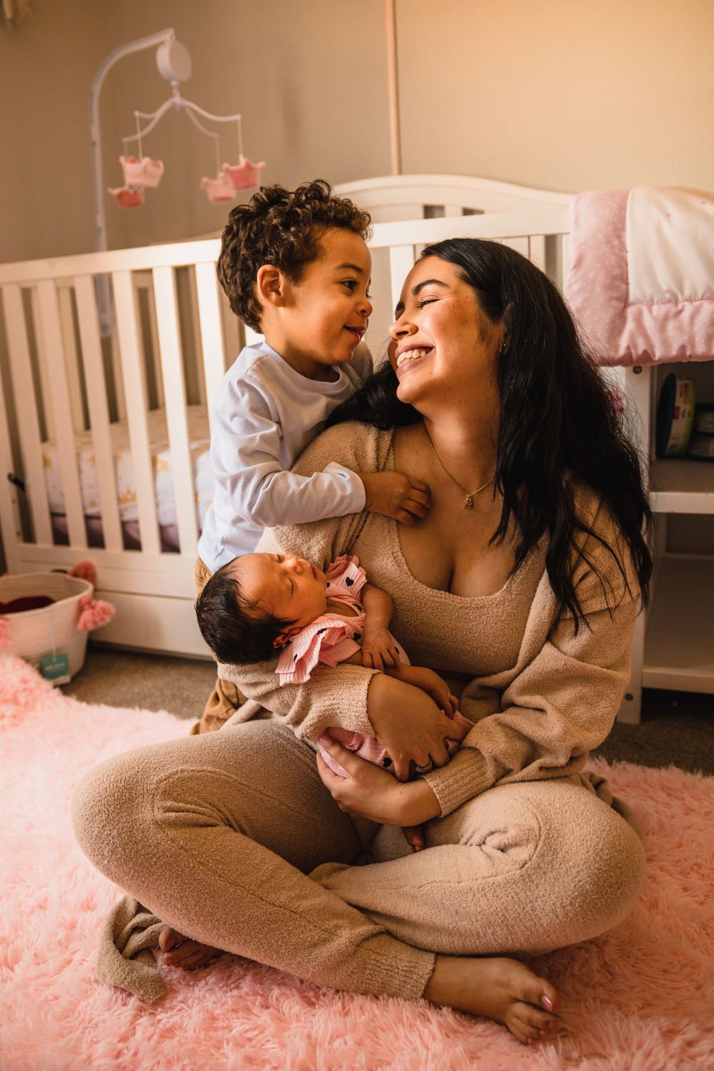 A joyful family moment on a pink rug as a toddler and parent share laughs while holding an infant.