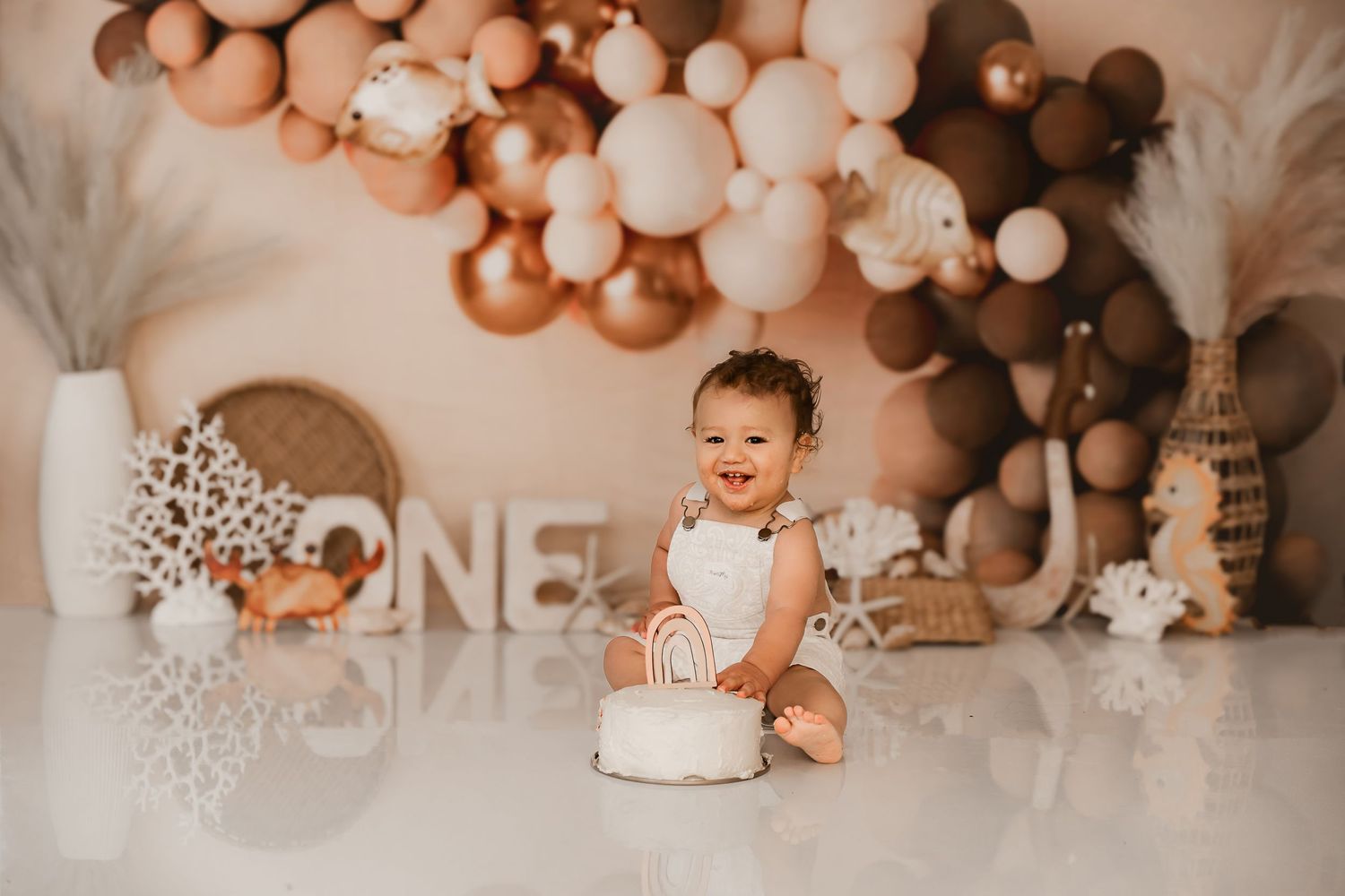 Baby first birthday cake smash photo with rose gold and white balloon arch and pampas grass decorations.