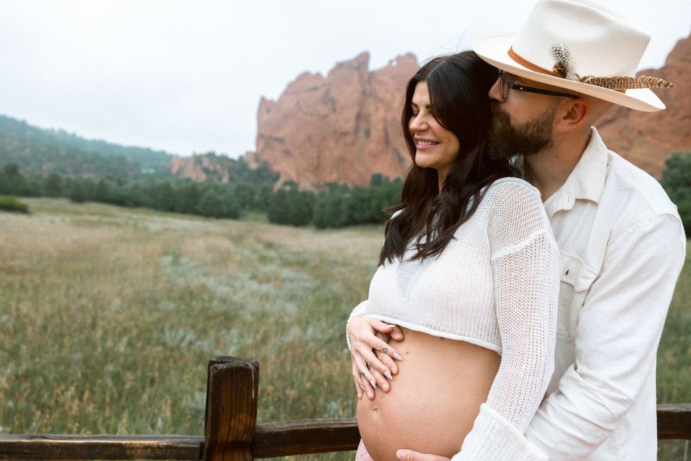 A pregnant couple in western attire stands in an open field with red rock formations visible in the background.