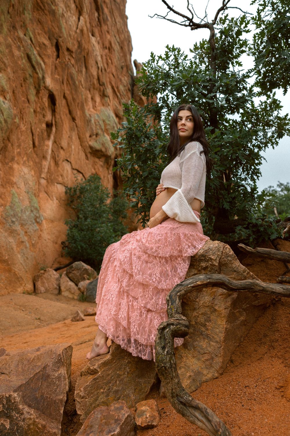 A model in a white sweater and pink ruffled maxi skirt poses on red rocks amid desert vegetation.