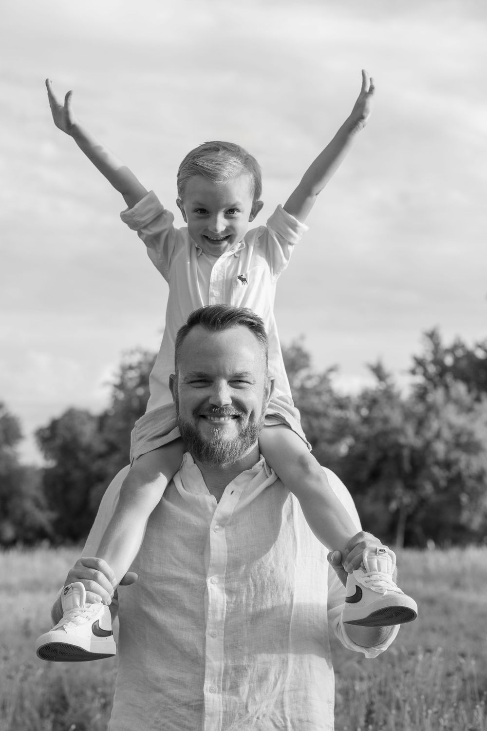 A joyful outdoor black and white portrait of a happy young child sitting on someone's shoulders with raised arms.