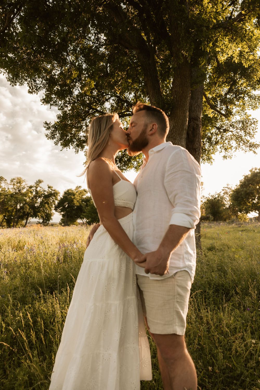 A couple shares a tender moment at sunset in a meadow under a large tree.
