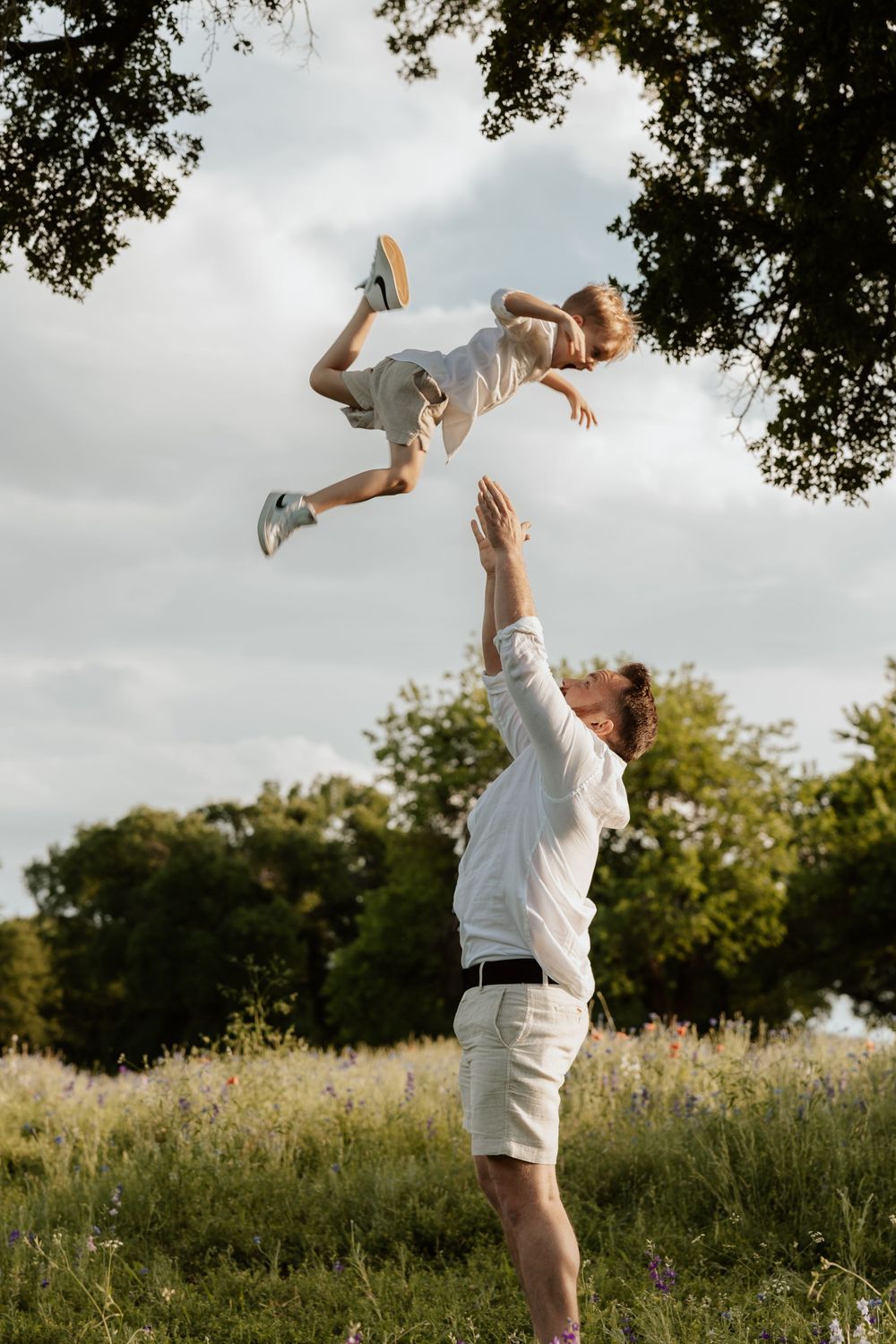 Joyful outdoor scene showing a child being playfully tossed into the air in a sunny meadow with trees in background.