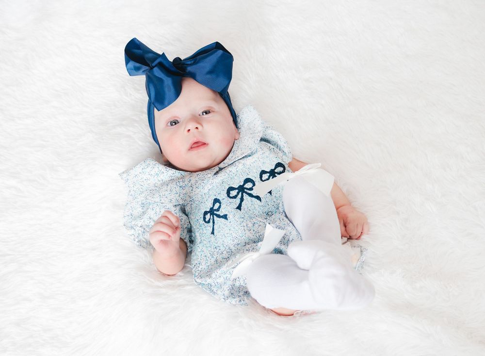 Baby wearing white outfit and blue bow headband lies on white bedding while looking at camera.