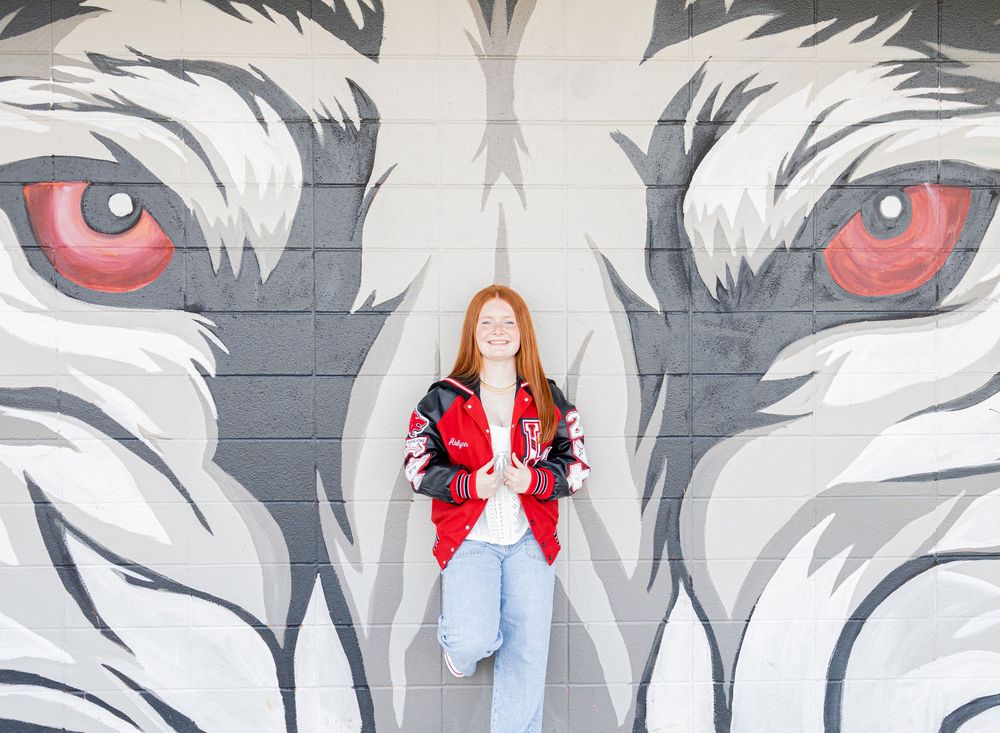 A person with red hair stands against a brick wall mural featuring large white and black wolf faces with red eyes.
