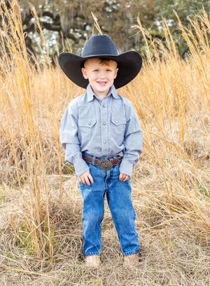 Young child wearing a cowboy hat, denim shirt, and jeans stands in tall golden grass during a sunny day.